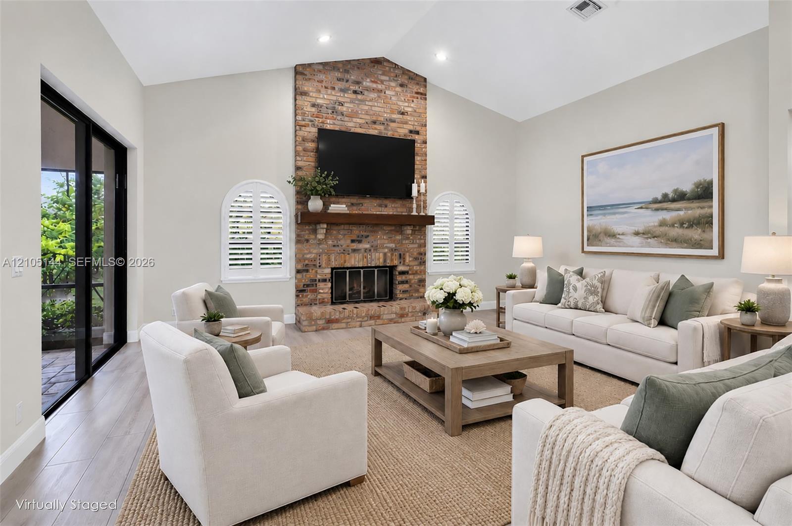 Living area featuring a floor-to-ceiling brick fireplace, natural wood mantel, vaulted ceilings, arched windows with plantation shutters, and wood-finish flooring