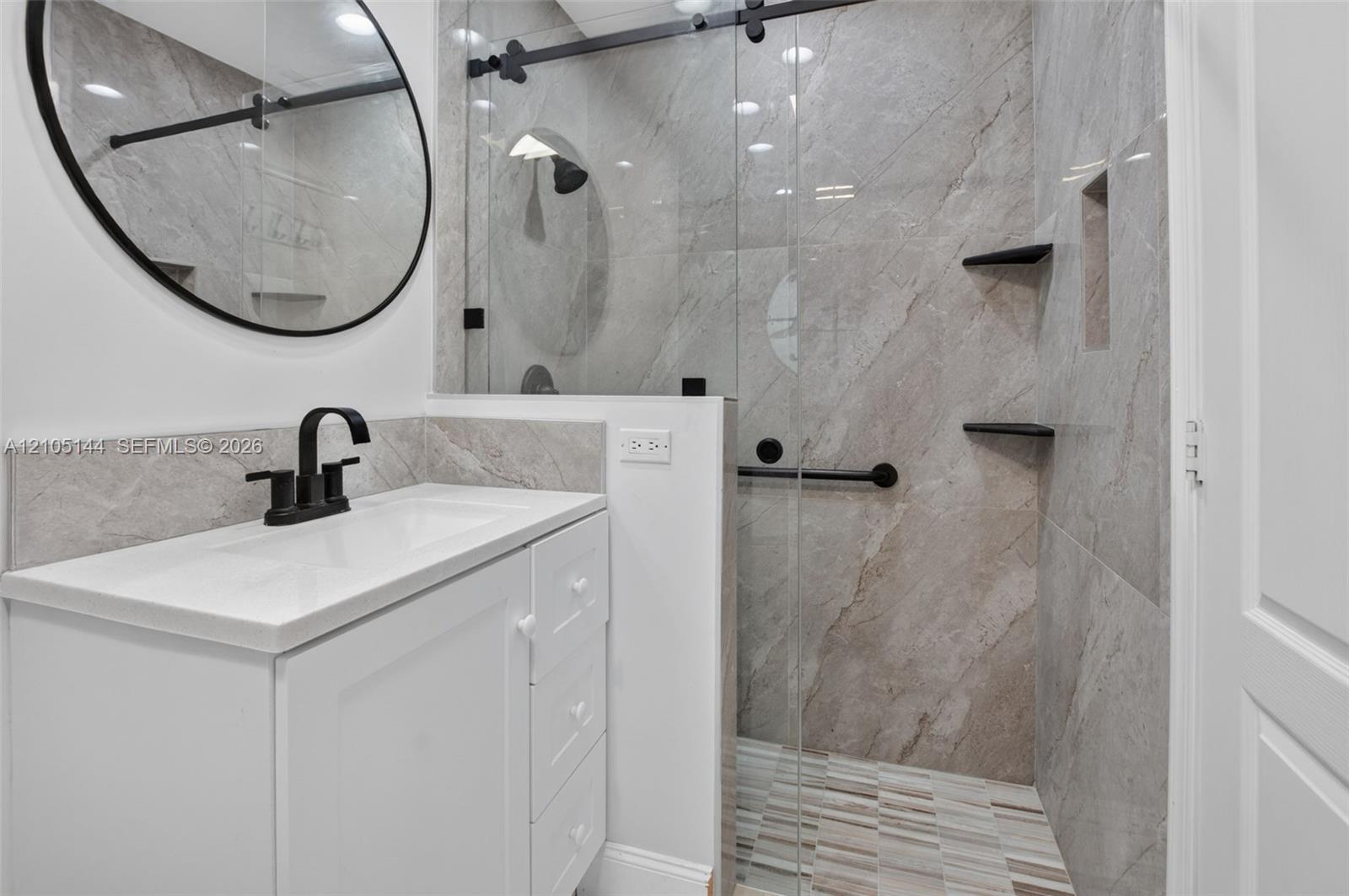 White vanity cabinet with an integrated rectangular sink and matte black faucet, paired with a frameless glass shower enclosure featuring large-format stone-tile walls, built-in shelving, and matte black hardware