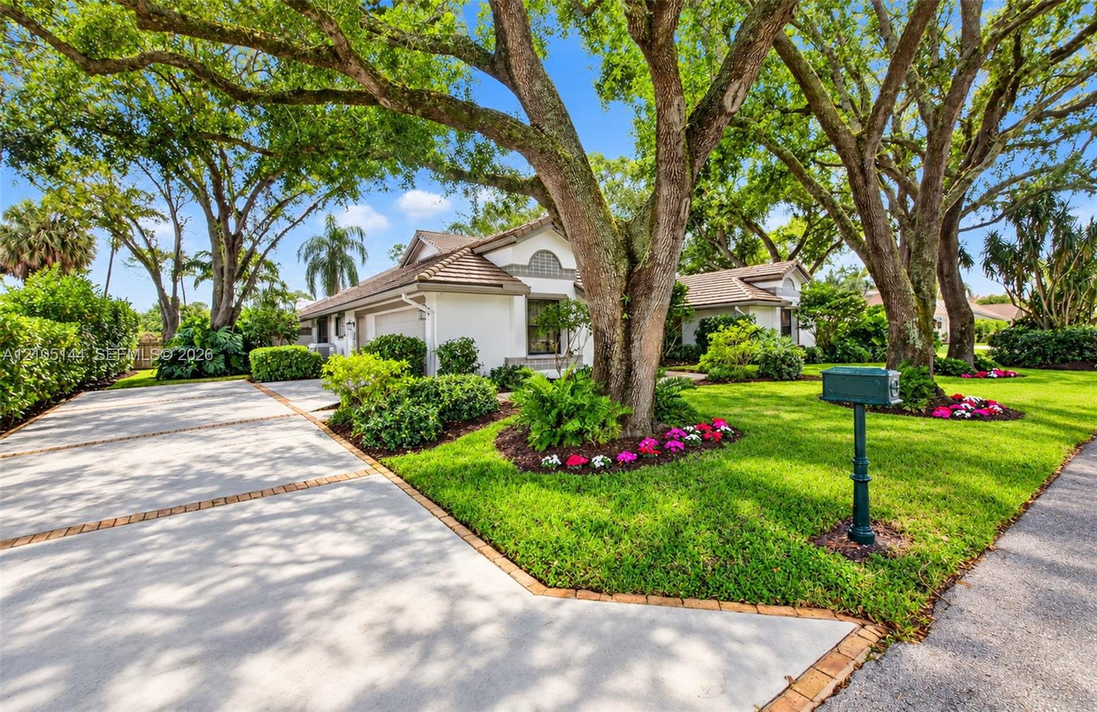 Stucco exterior residence featuring a concrete driveway with brick paver borders, mature canopy trees, and landscaped garden beds