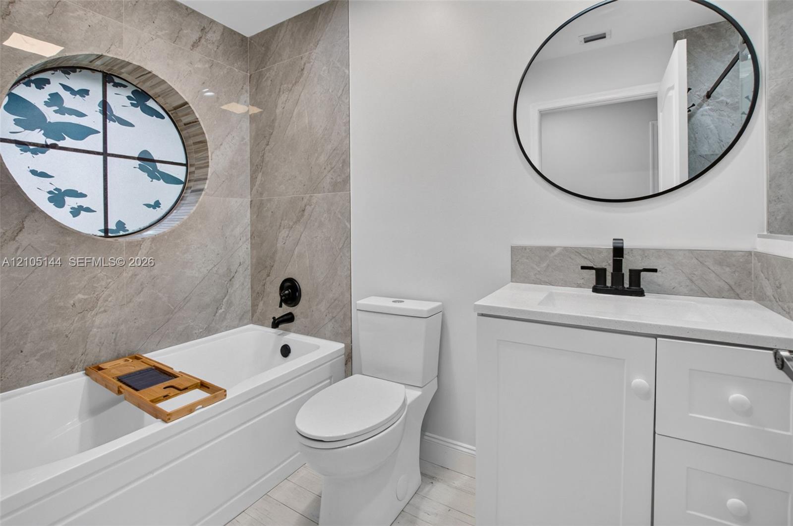 Bathroom featuring large-format tile surround, circular accent window, white vanity with integrated basin, matte black fixtures, and oval wall-mounted mirror