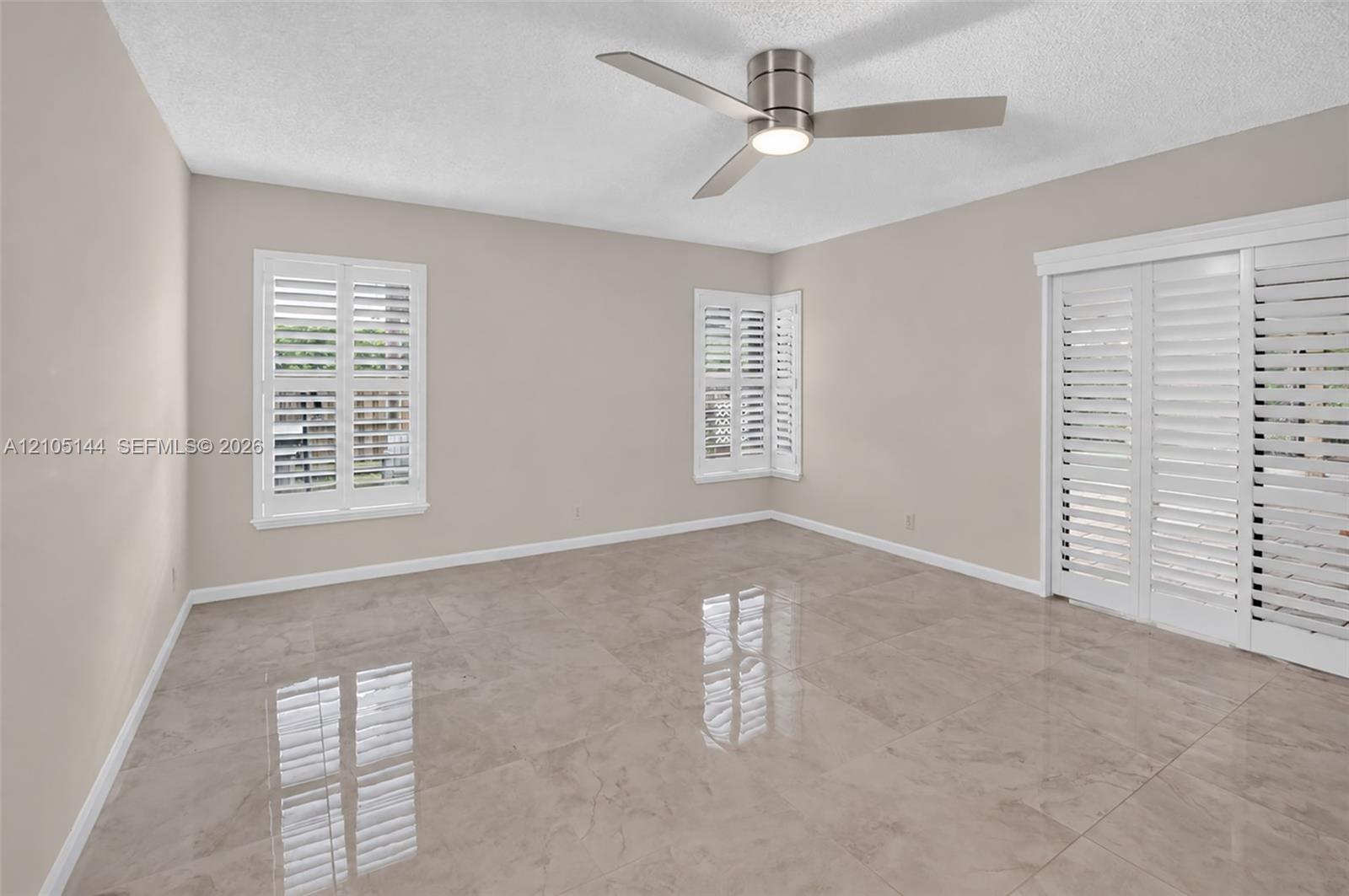 (FAN VIRTUALLY STAGED AS SIMILAR PIECE WILL BE INSTALLED)Large-format neutral tile flooring, triple white plantation shutters, and a modern brushed-nickel ceiling fan with integrated lighting