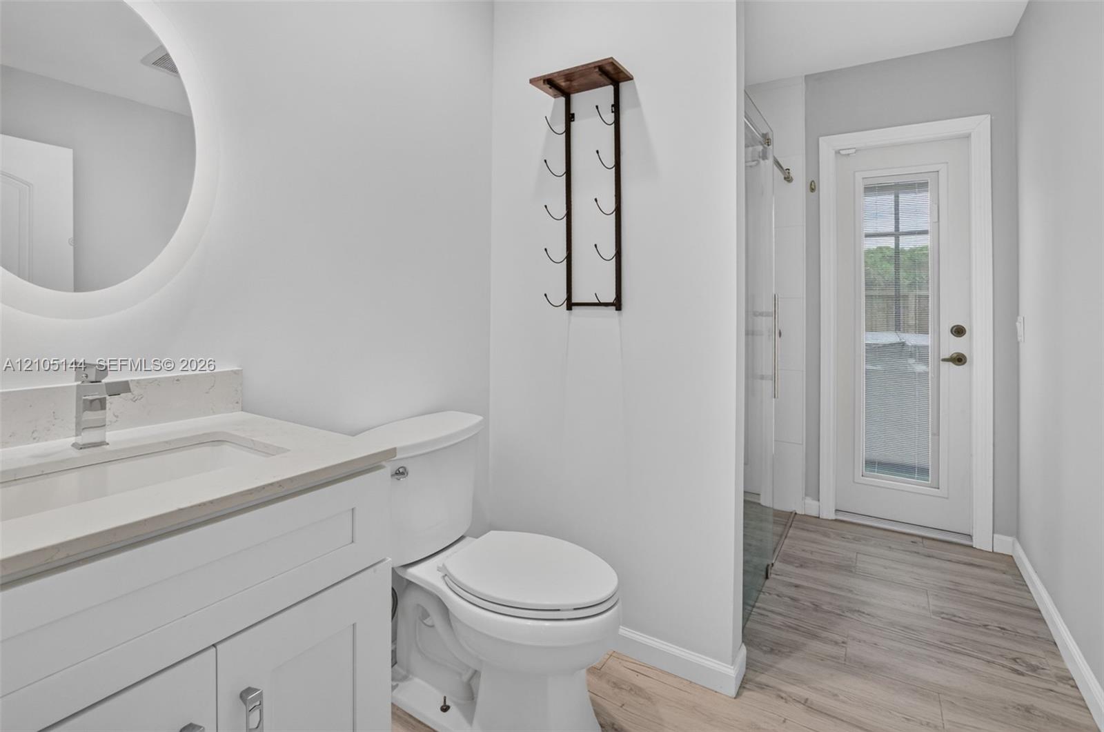 Bathroom featuring a white vanity with a light-toned stone countertop, integrated sink with a chrome faucet, circular LED-backlit mirror, and wood-finish flooring