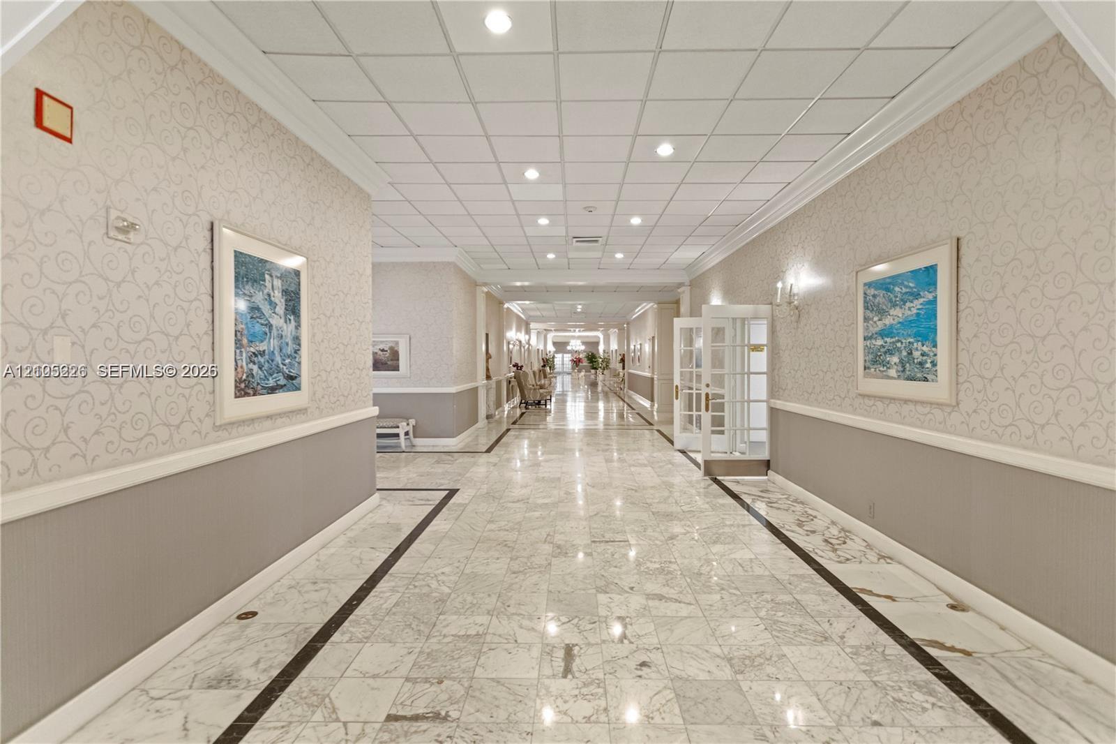 Spacious corridor featuring polished stone flooring with decorative inlay, recessed grid ceiling, custom wall molding, and integrated wall sconces