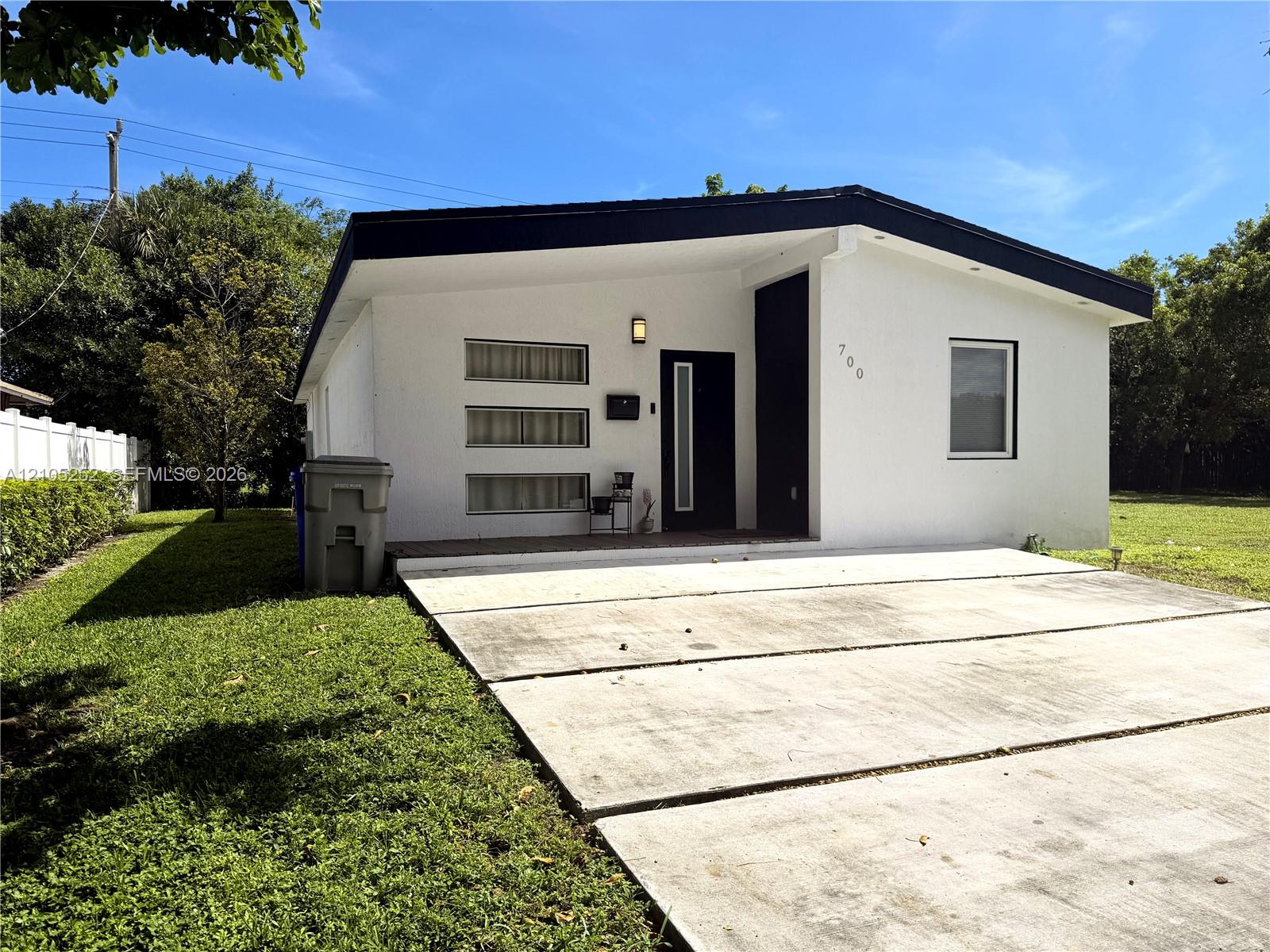 White stucco exterior with black trim, modern entry door, concrete panel driveway, and a white fence