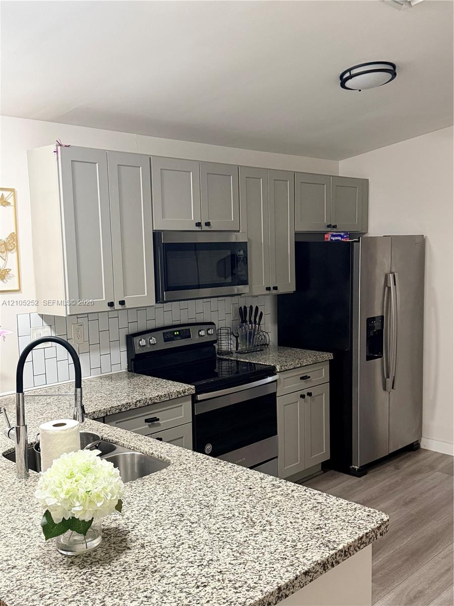 Updated kitchen featuring granite countertops, stainless steel appliances, light gray shaker cabinetry, a subway tile backsplash, and wood-finish flooring