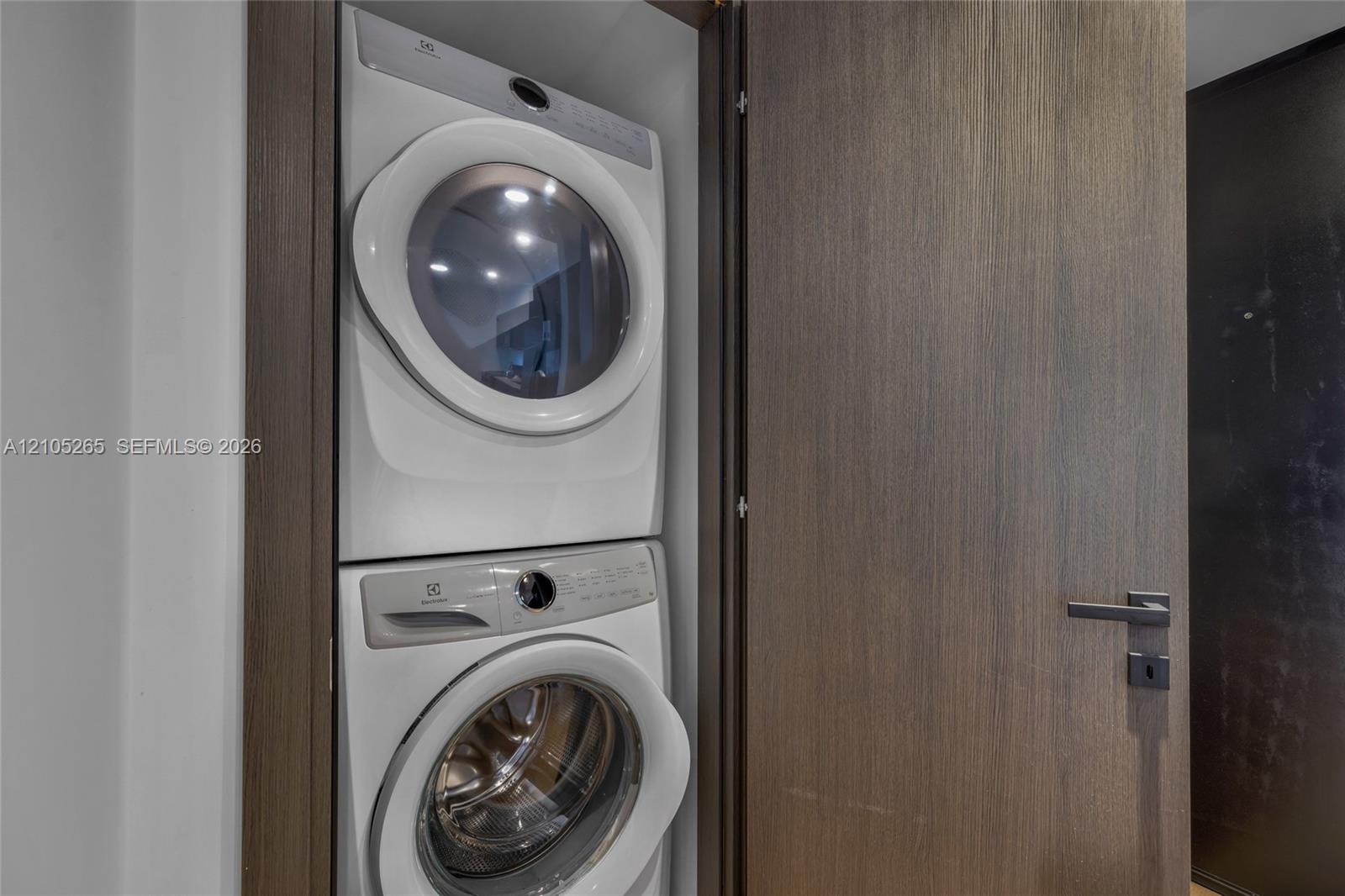 Stacked white front-load washer and dryer unit enclosed in a closet with a full-height, dark wood-finish door and integrated metal lever handle