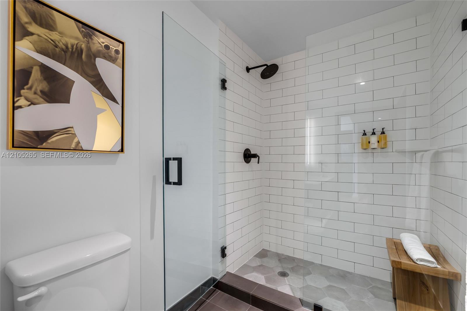 Walk-in shower with white subway tile surround, matte black rainfall shower head, hexagon floor tile, glass door, and built-in stone corner shelf