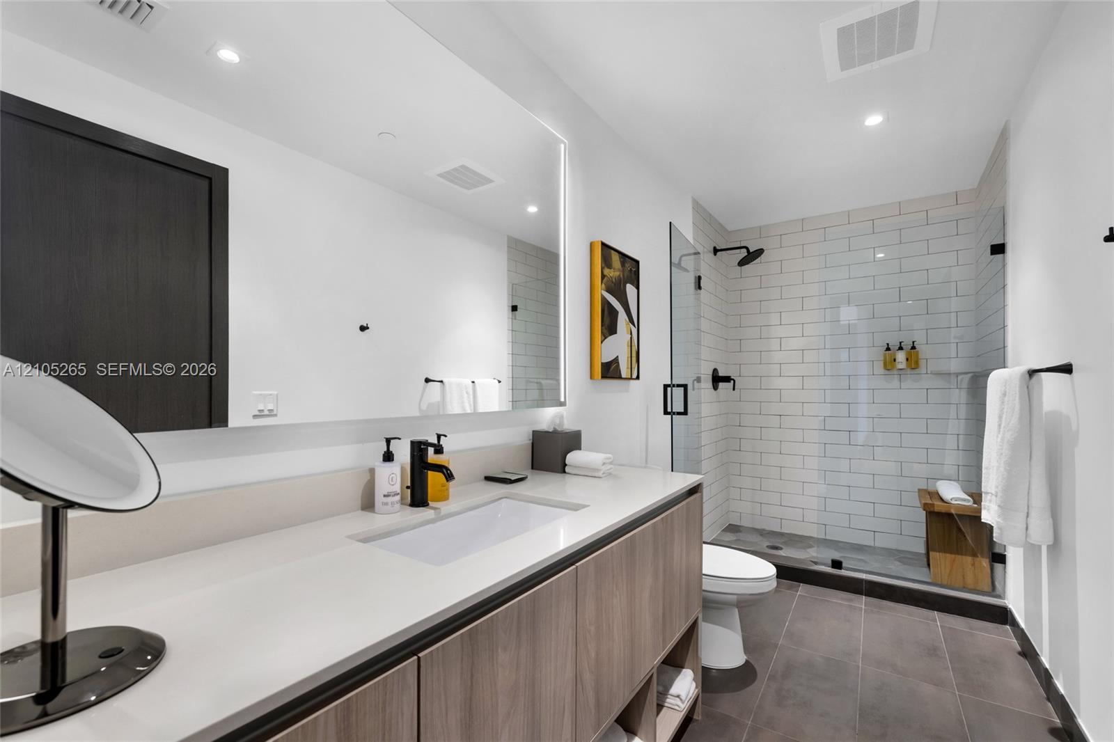 Floating wood-finish vanity with integrated sink and quartz countertop, large backlit mirror, and glass-enclosed shower featuring white subway-style tile and matte black fixtures