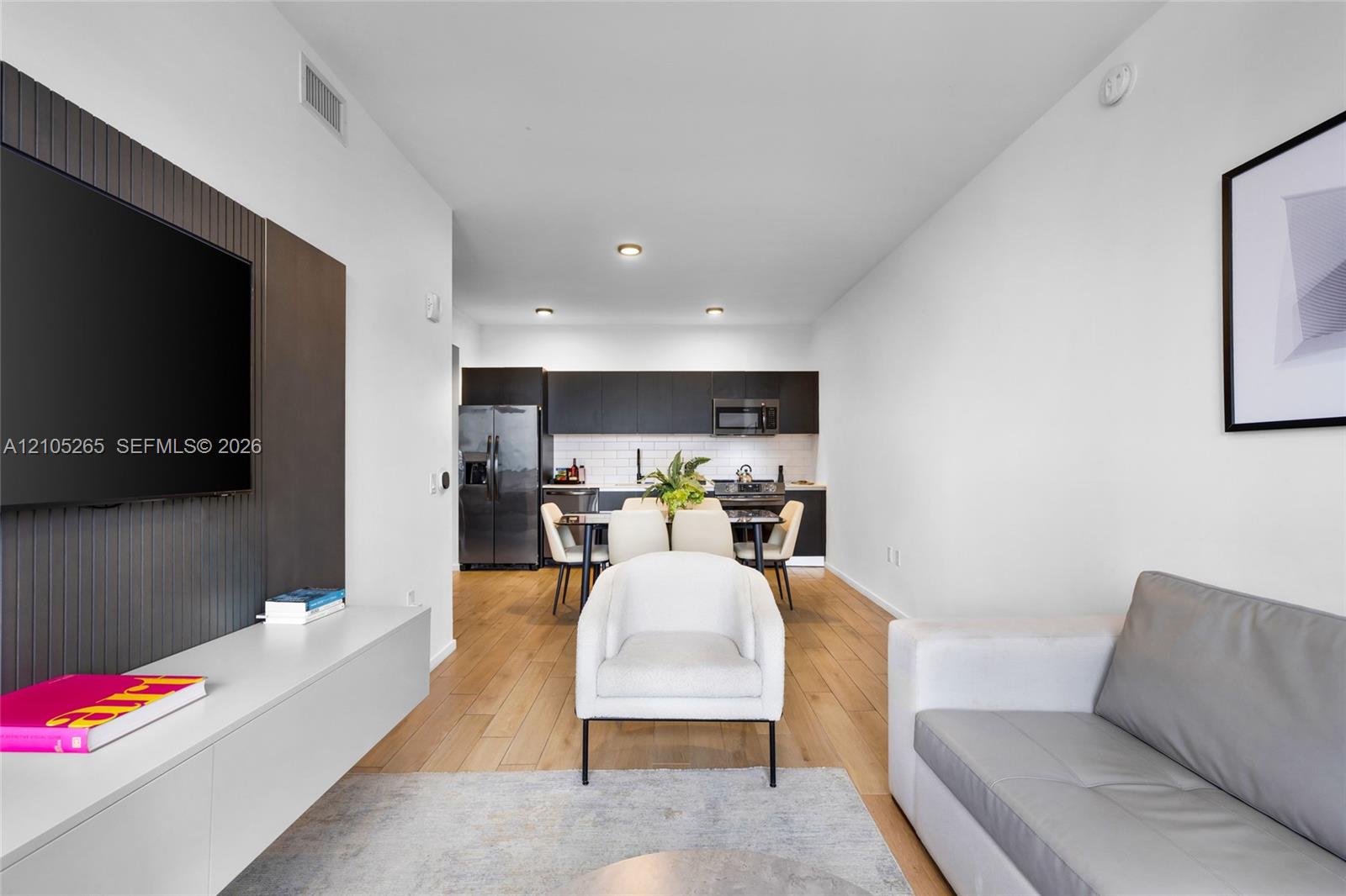 Modern interior featuring wood-finish flooring, a matte-finish accent wall with a wall-mounted media console, dark cabinetry, and white tile backsplash