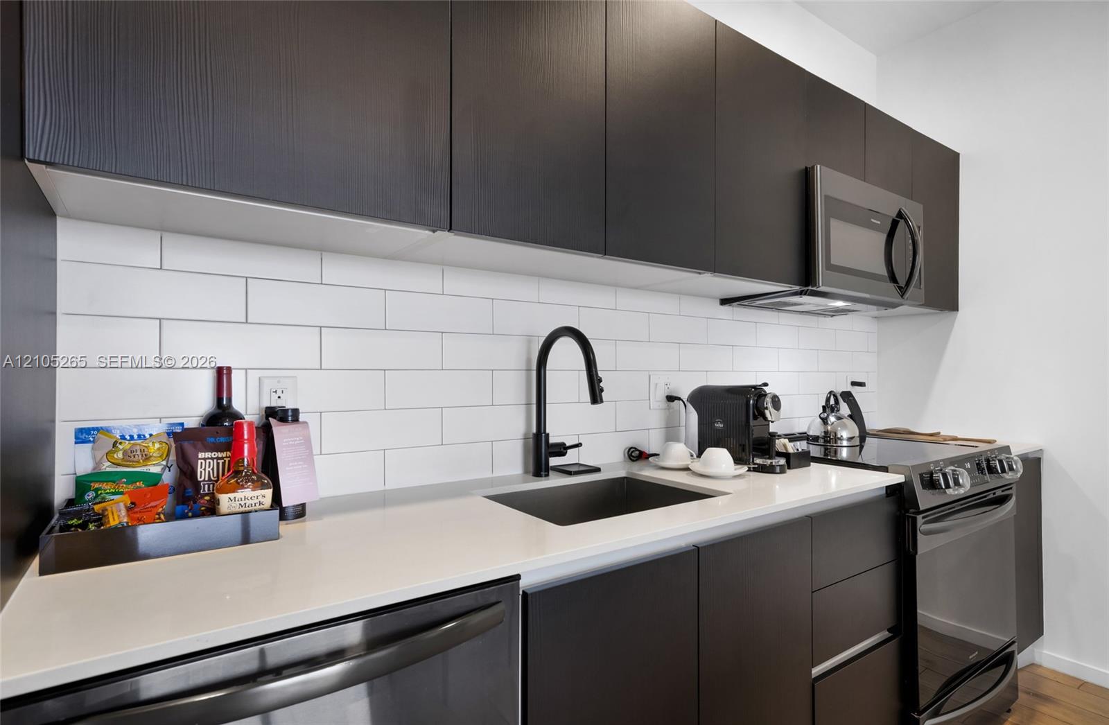 Modern kitchen featuring dark slab-style cabinetry, light-toned countertops, white subway tile backsplash, matte black hardware, and stainless steel appliances