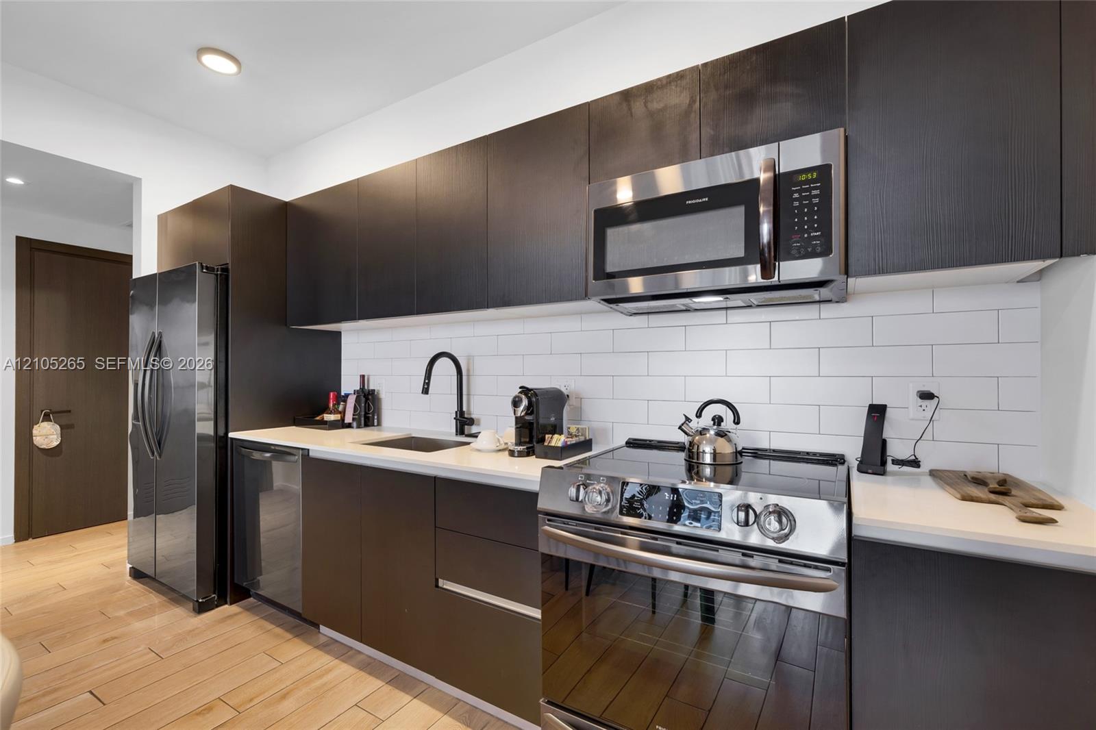 Modern kitchen featuring dark cabinetry, white subway tile backsplash, solid-surface countertops, stainless steel appliances, and wood-finish flooring