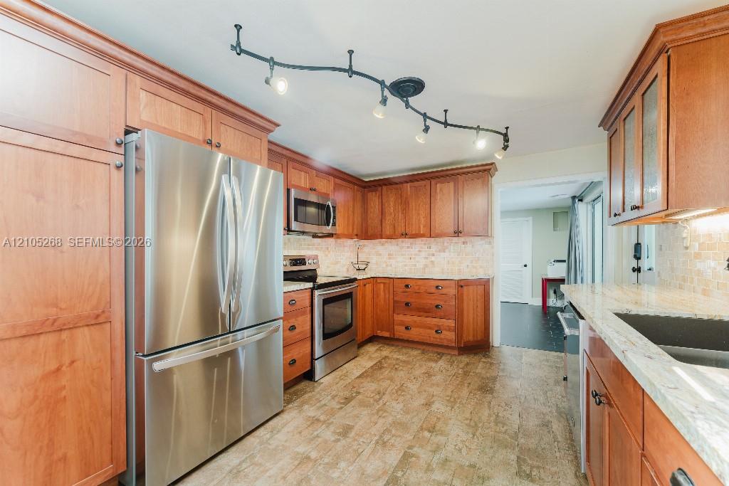 Kitchen featuring warm wood cabinetry, stainless steel appliances, light-toned backsplash, granite countertops, and track lighting