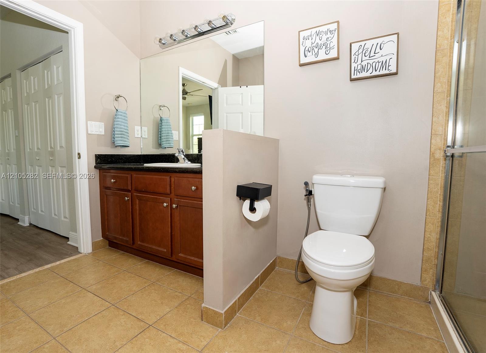 Bathroom featuring wood-finish cabinetry, dark stone-surfaced vanity top, large wall-mounted mirror, multi-bulb light fixture, and tile flooring