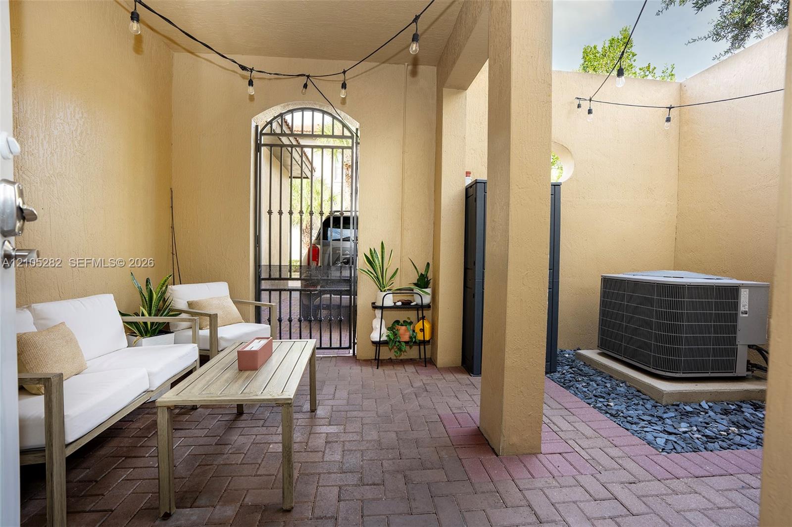 Covered patio featuring red brick paver flooring, a decorative arched wrought-iron gate, and built-in string lighting