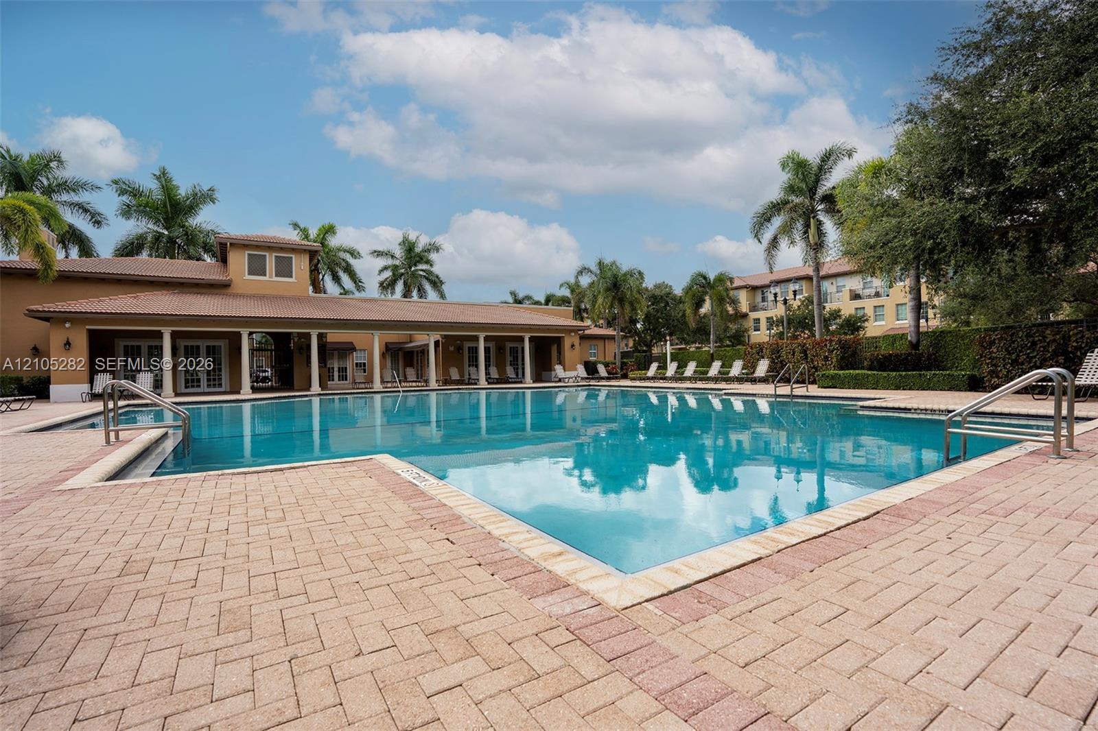 Outdoor swimming pool featuring a brick-paved deck, covered veranda with structural columns, and surrounding palm trees