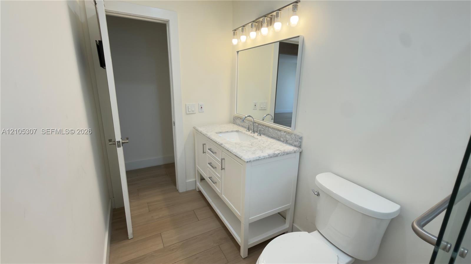 Bathroom vanity featuring stone-finish countertop, integrated sink, and white cabinetry, accompanied by a rectangular mirror, five-light fixture, and wood-finish flooring Bathroom vanity featuring stone-finish countertop, integrated sink, and white cabinetry, accompanied by a rectangular mirror, five-light fixture, and wood-finish flooring
