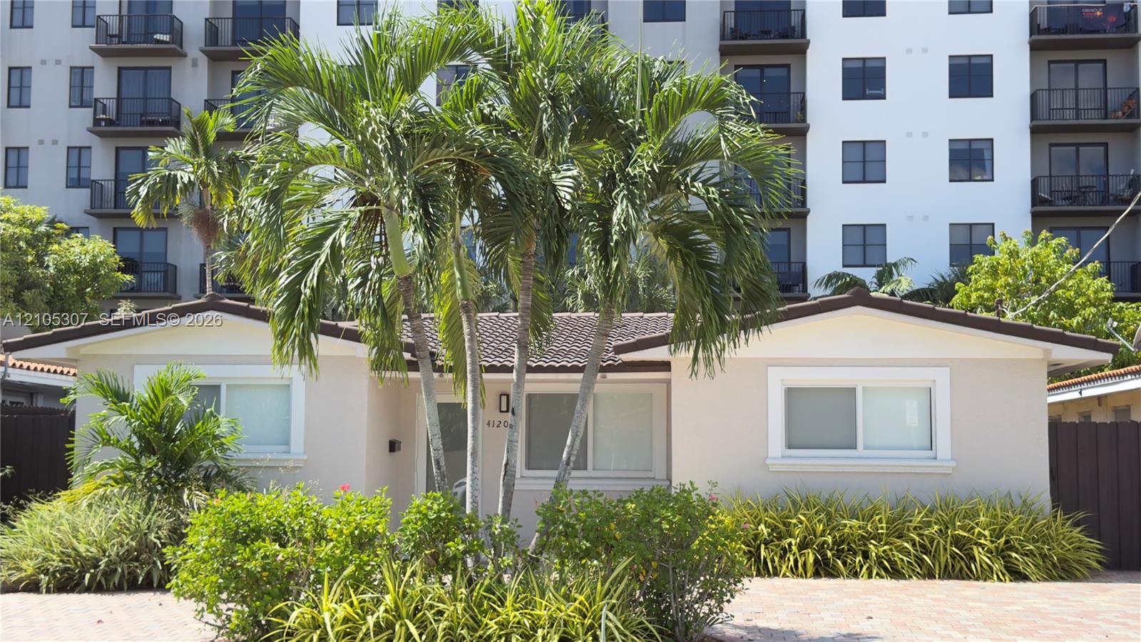 Single-story exterior featuring a light-colored stucco facade, tiled roof, paver driveway, and mature tropical landscaping Single-story exterior featuring a light-colored stucco facade, tiled roof, paver driveway, and mature tropical landscaping