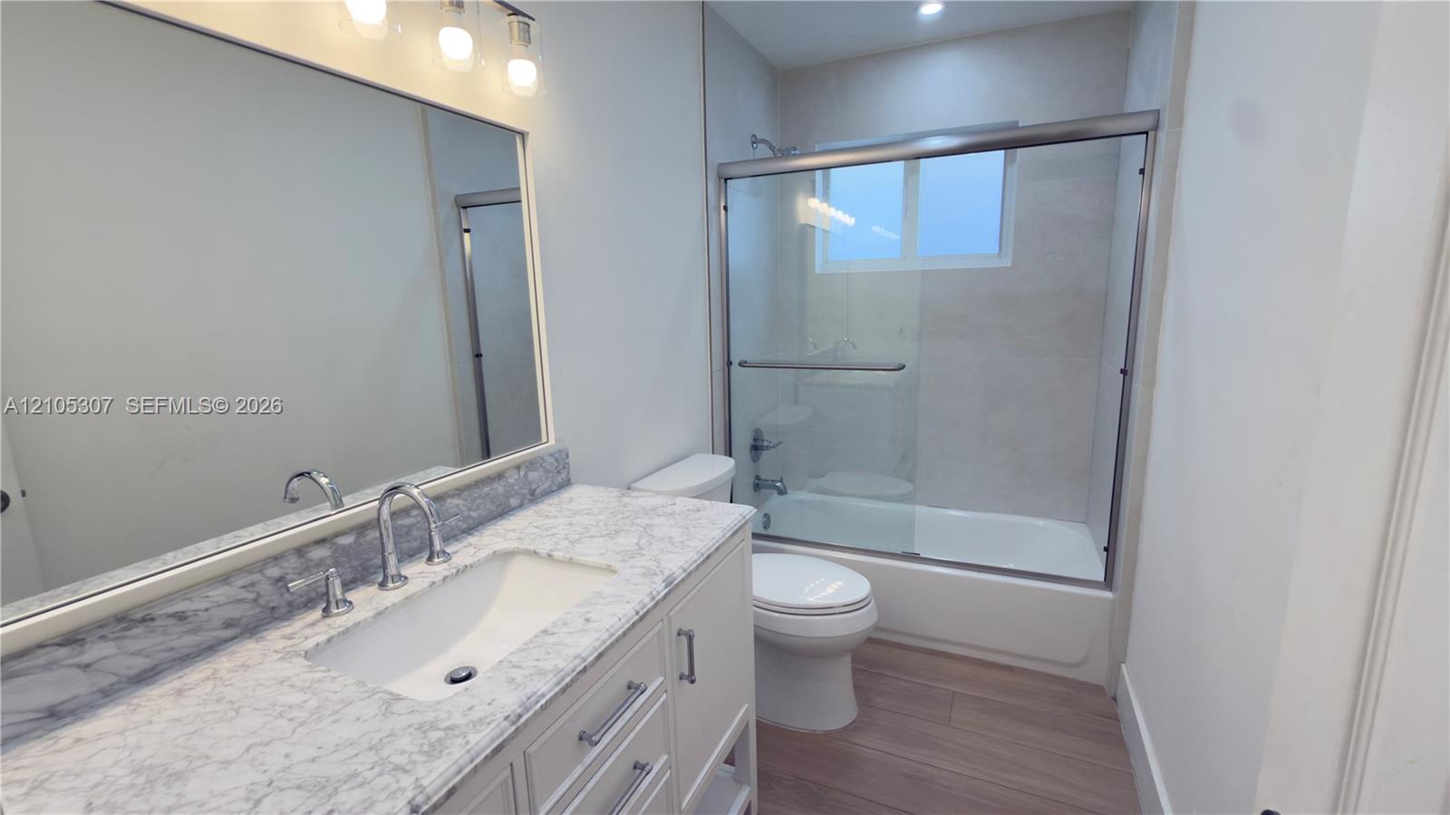Bathroom with tub, eaturing a vanity with a stone-finish countertop, undermount sink, chrome faucet, large wall-mounted mirror with integrated lighting, and a tub-shower combination with a glass sliding enclosure Bathroom with tub, eaturing a vanity with a stone-finish countertop, undermount sink, chrome faucet, large wall-mounted mirror with integrated lighting, and a tub-shower combination with a glass sliding enclosure
