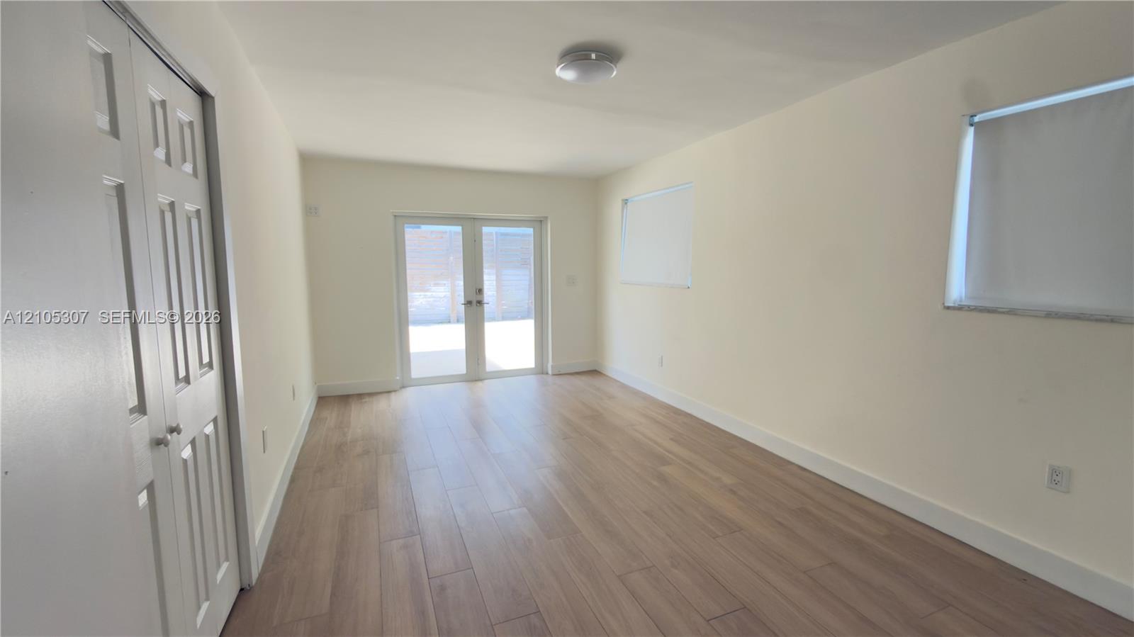 Interior space featuring light-toned wood-finish flooring, dual-panel closet doors, white walls, and French doors leading to a paved patio area Interior space featuring light-toned wood-finish flooring, dual-panel closet doors, white walls, and French doors leading to a paved patio area