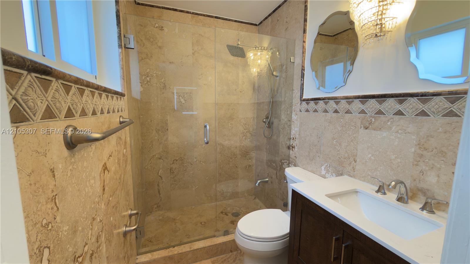 Bathroom featuring natural stone wall tile with decorative accent border, a glass-enclosed shower with a rainfall showerhead, and a vanity with a white rectangular sink and countertop Bathroom featuring natural stone wall tile with decorative accent border, a glass-enclosed shower with a rainfall showerhead, and a vanity with a white rectangular sink and countertop