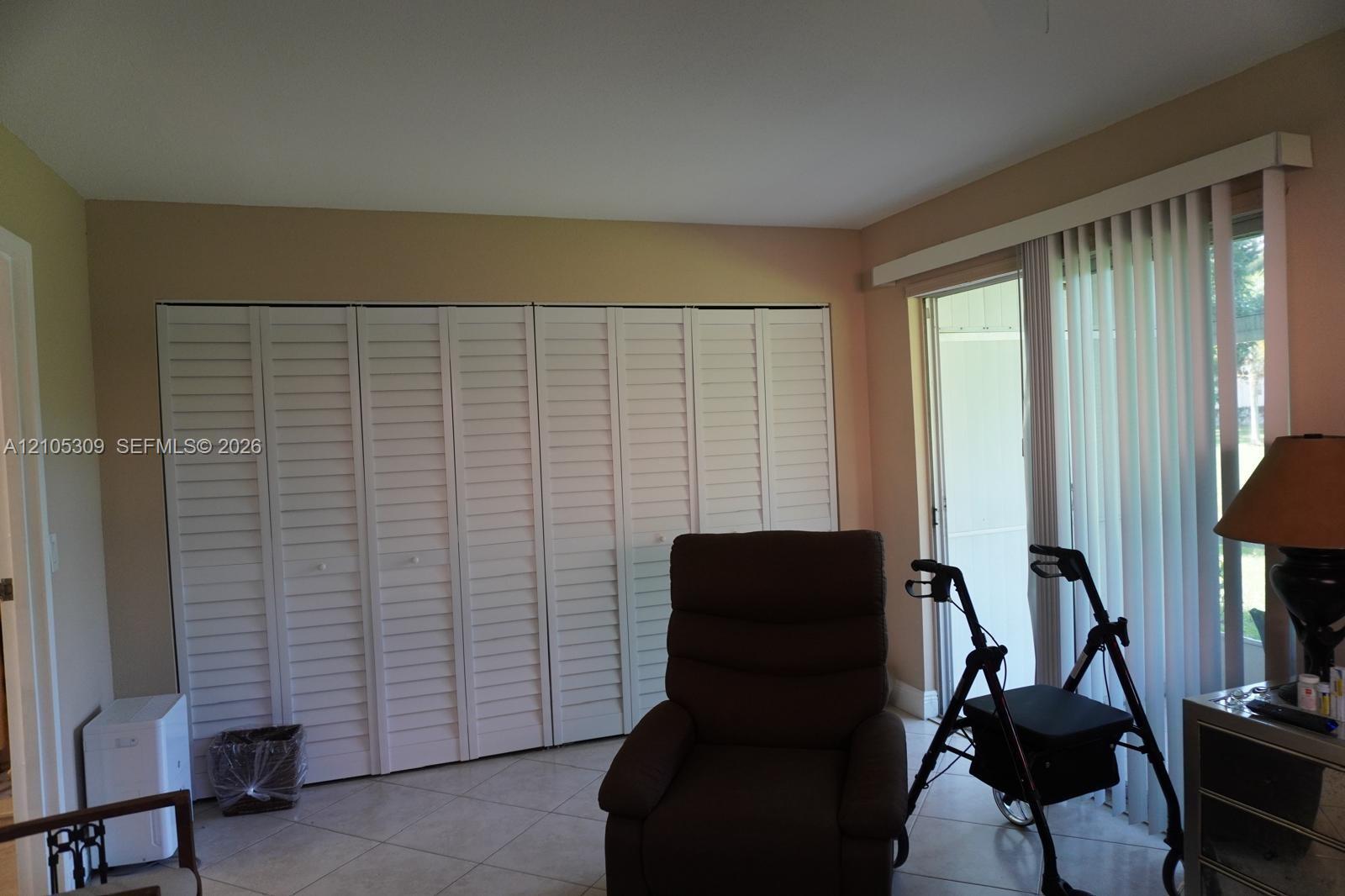 Full-wall louvered closet doors, neutral wall finish, ceramic tile flooring, and vertical blinds covering the sliding glass door Full-wall louvered closet doors, neutral wall finish, ceramic tile flooring, and vertical blinds covering the sliding glass door