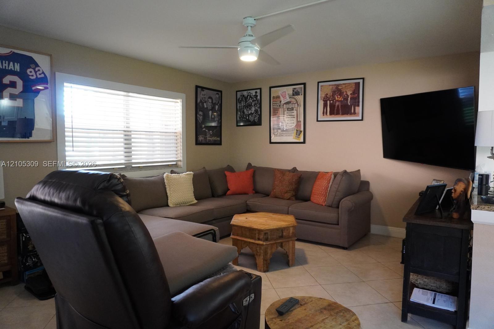 Neutral interior featuring light-toned tile flooring, a dual-function ceiling fan with integrated lighting, and a large picture window with horizontal blinds Neutral interior featuring light-toned tile flooring, a dual-function ceiling fan with integrated lighting, and a large picture window with horizontal blinds