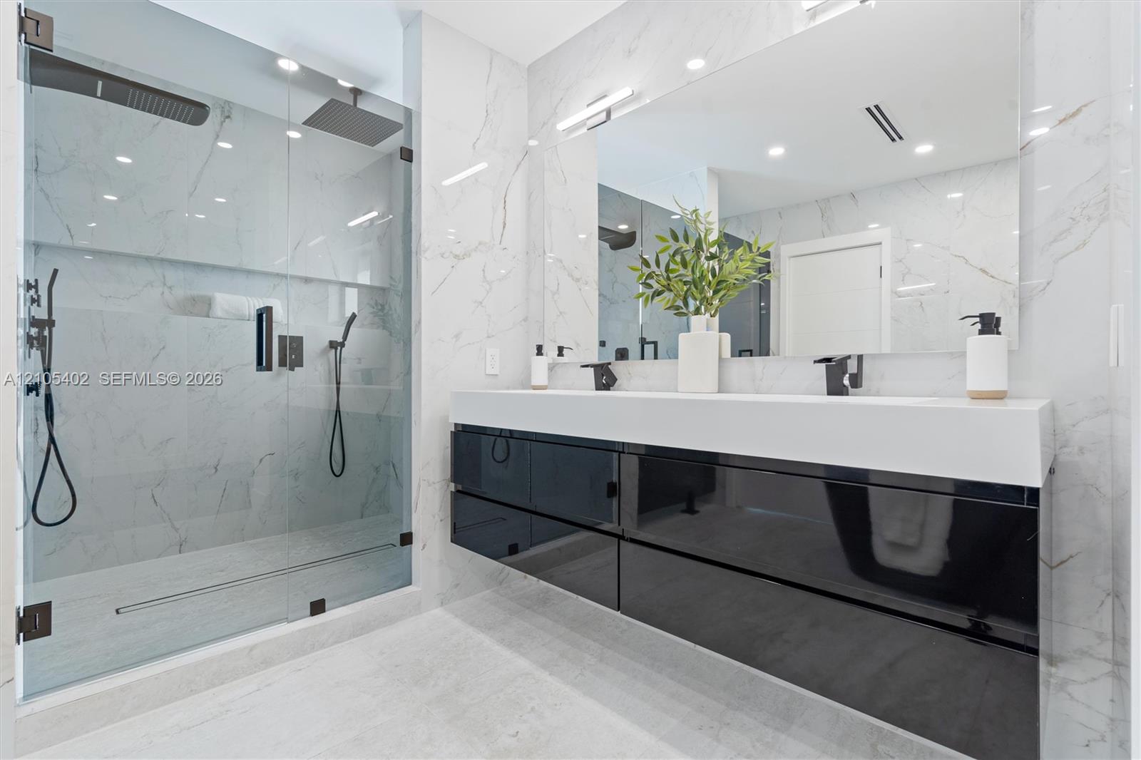 Floating high-gloss black vanity featuring a dual-sink white countertop, wall-to-wall mirror, glass-enclosed walk-in shower with stone-finish wall tile, and matte black fixtures
