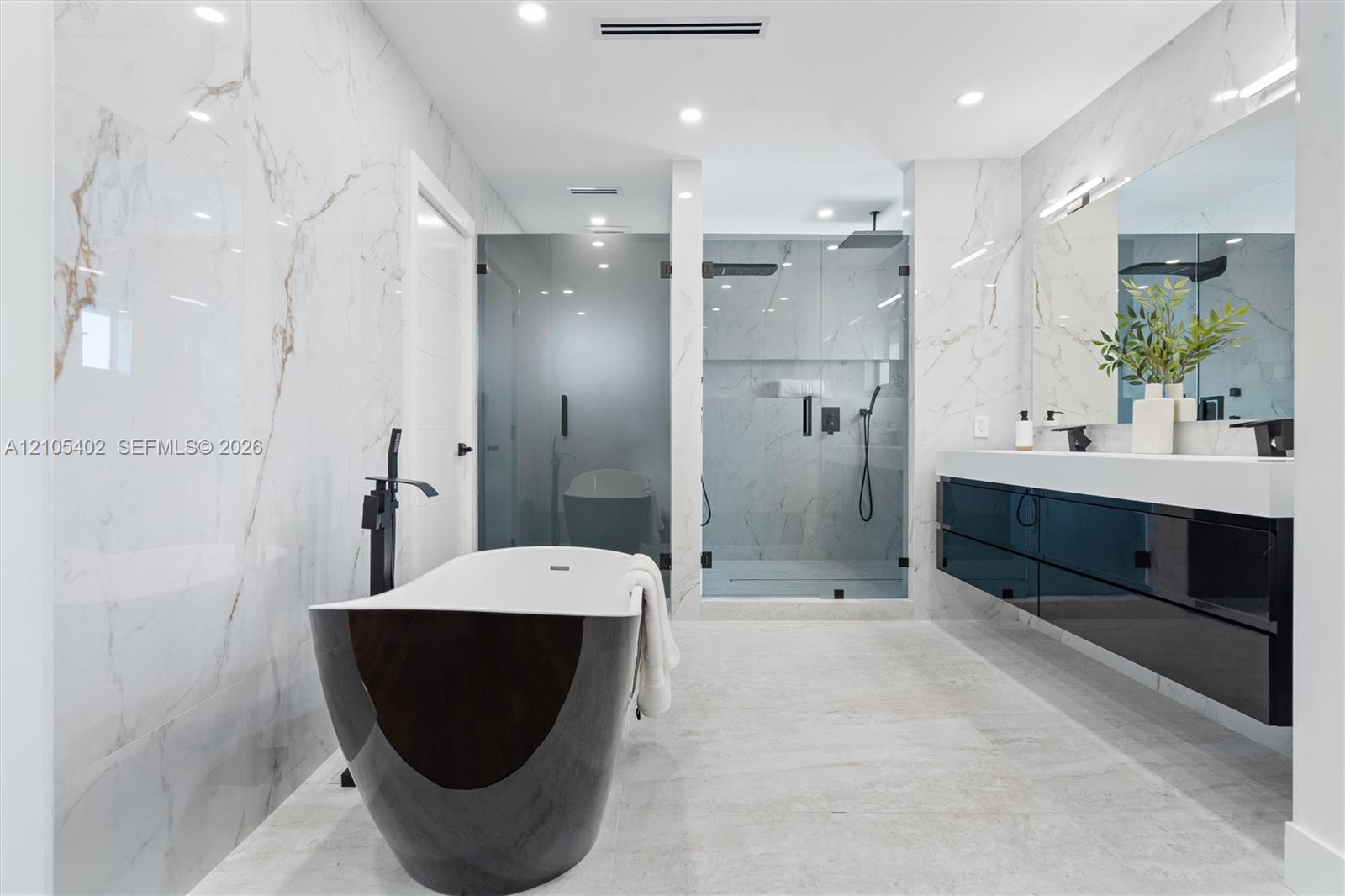Freestanding deep-soaking tub with floor-mounted matte black faucet, wall-to-wall stone-finish tile surround, wall-mounted floating dual vanity with integrated countertop, and walk-in glass-enclosed shower with matte black fixtures