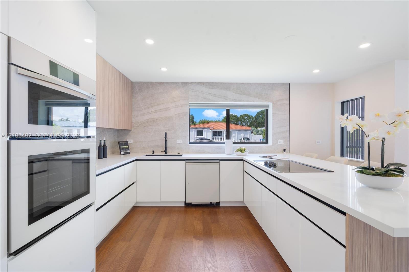 Modern kitchen featuring white flat-panel cabinetry, wood-finish cabinetry accents, quartz countertops, a stone-patterned backsplash, and integrated stainless steel appliances