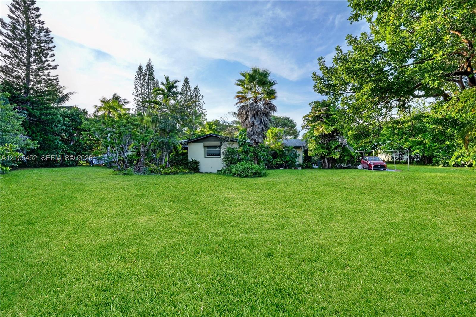 Expansive lawn featuring mature trees, diverse tropical landscaping, and a detached carport structure