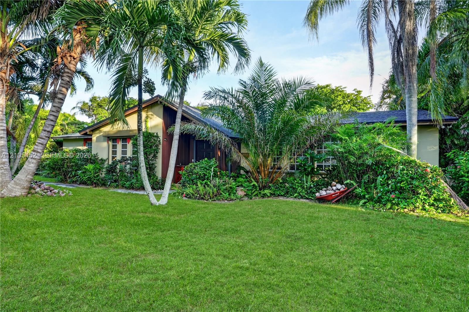 Single-story exterior featuring a low-pitch roofline, painted siding, mature palm trees, and extensive tropical landscaping