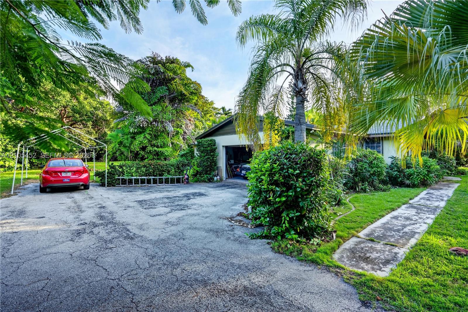 Asphalt driveway featuring an attached garage and a concrete walkway, complemented by mature landscaping and established palm trees
