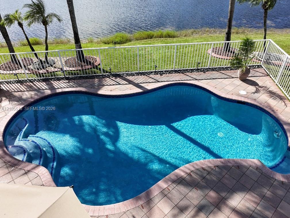 Freeform swimming pool with integrated steps, surrounded by paver decking, and secured by a white perimeter fence