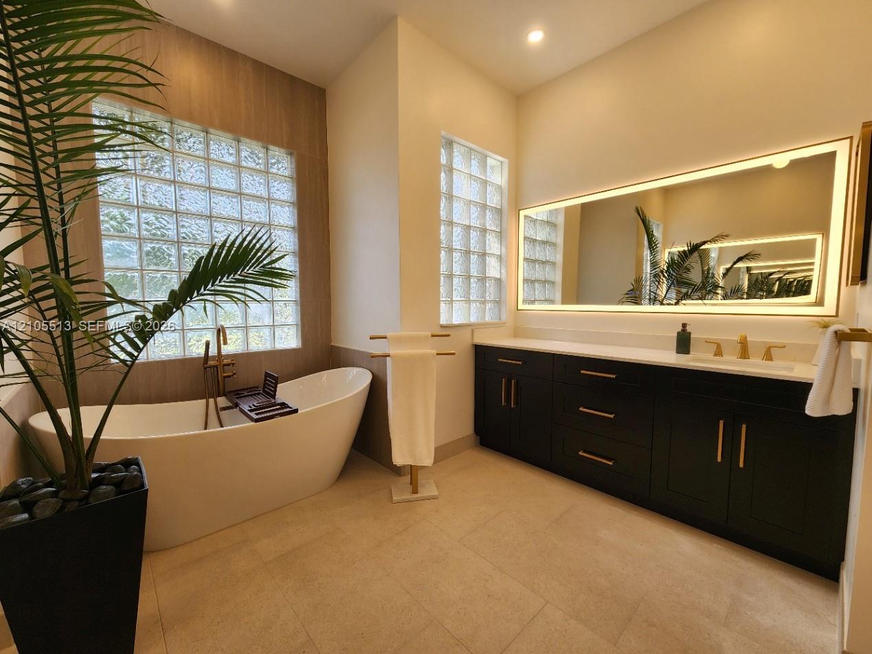 Spacious newly remodeled spa-inspired primary bathroom featuring porcelain tile, a freestanding soaking tub, dual custom vanities with quartz counters, recessed lighting, glass block windows, lighted mirrors and a tray ceiling with a modern chandelier