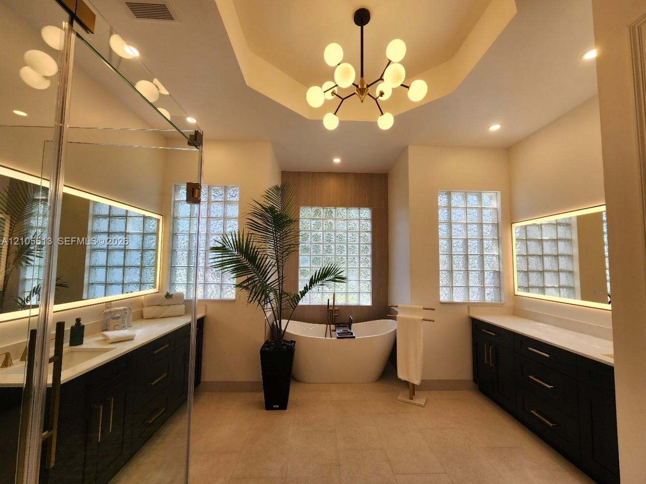 Spacious newly remodeled spa-inspired primary bathroom featuring porcelain tile, a freestanding soaking tub, dual custom vanities with quartz counters, recessed lighting, glass block windows, lighted mirrors and a tray ceiling with a modern chandelier