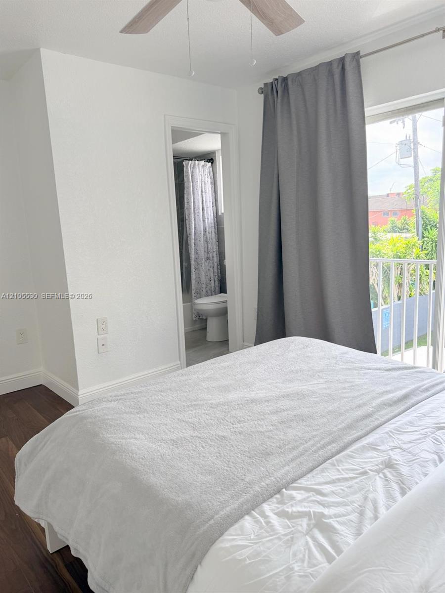 Wood-finish flooring, ceiling fan, sliding glass door, white balcony railing, and a bathroom entrance with a toilet and shower curtain