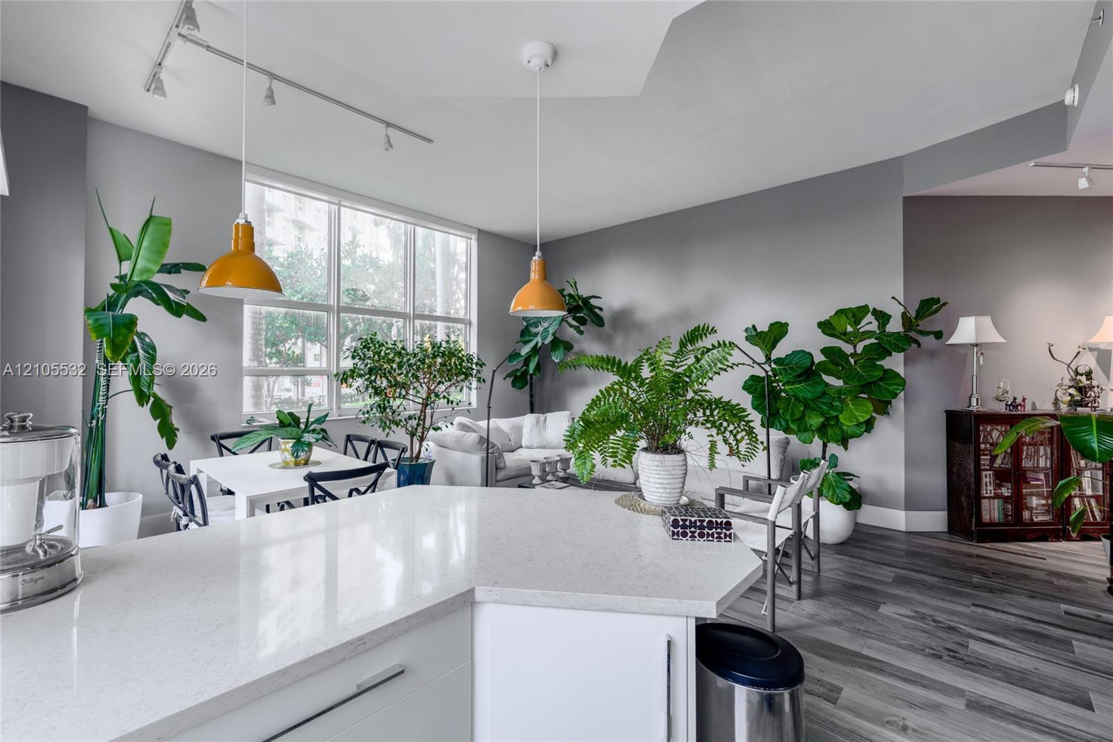 Open-concept interior featuring light-toned countertops, wood-finish flooring, recessed tray ceiling, multi-pane floor-to-ceiling windows, and contemporary pendant lighting