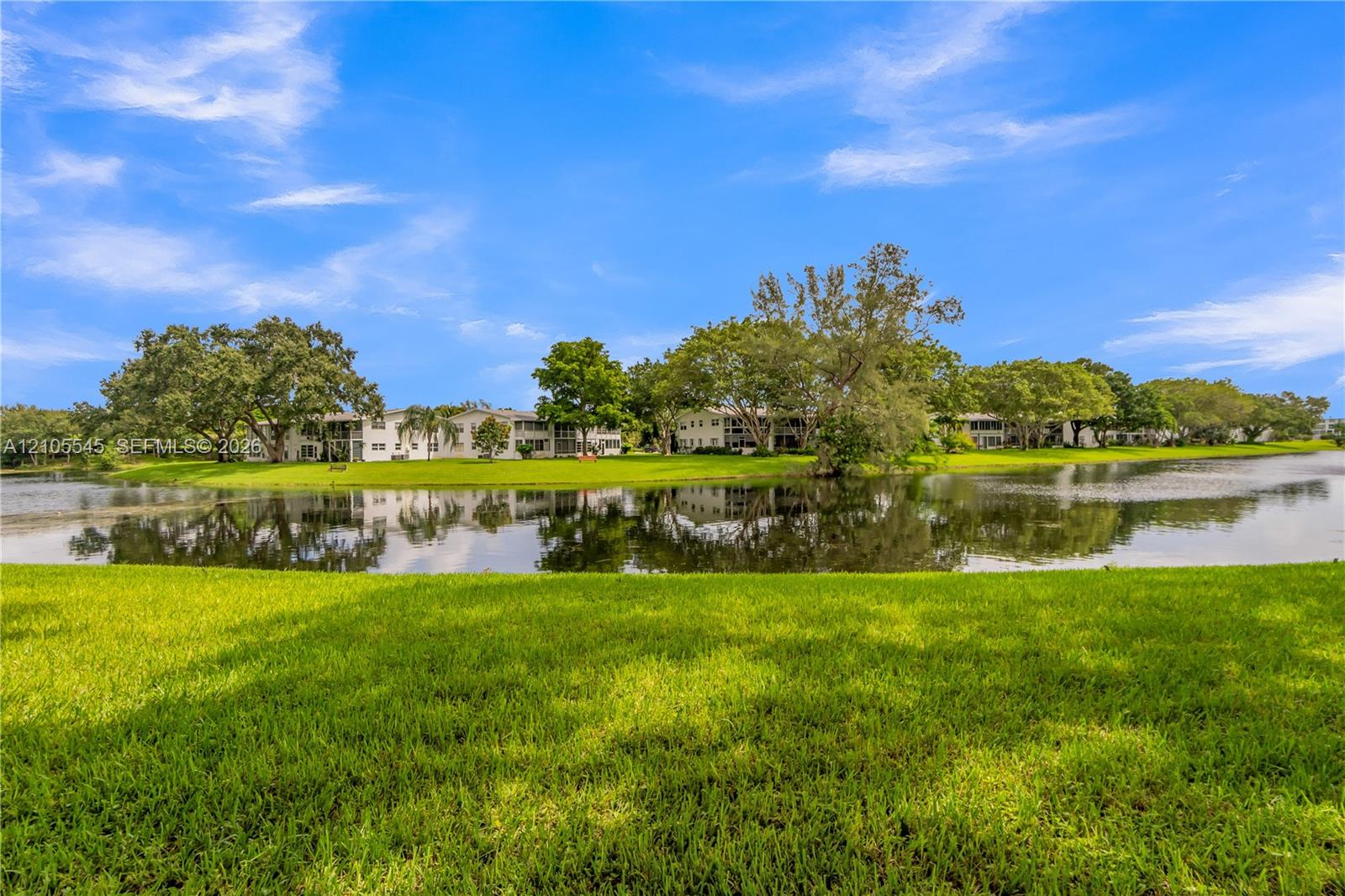 Expansive green lawn bordering a reflective body of water with established trees and multiple residential buildings in the background