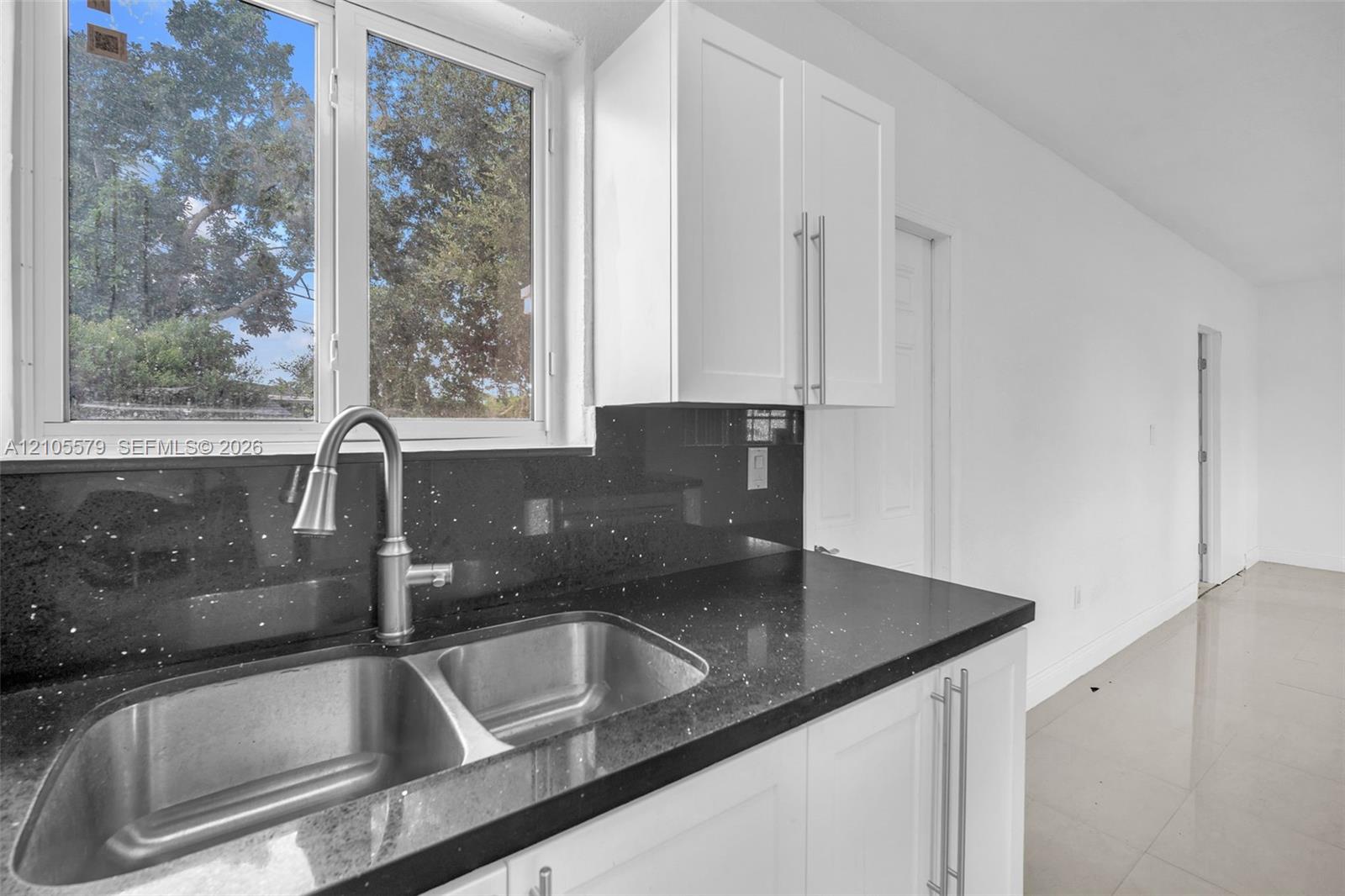 Kitchen area featuring white shaker-style cabinetry, dark stone-finish countertops, stainless steel dual-basin sink, high-arc brushed nickel faucet, and large-format tile flooring