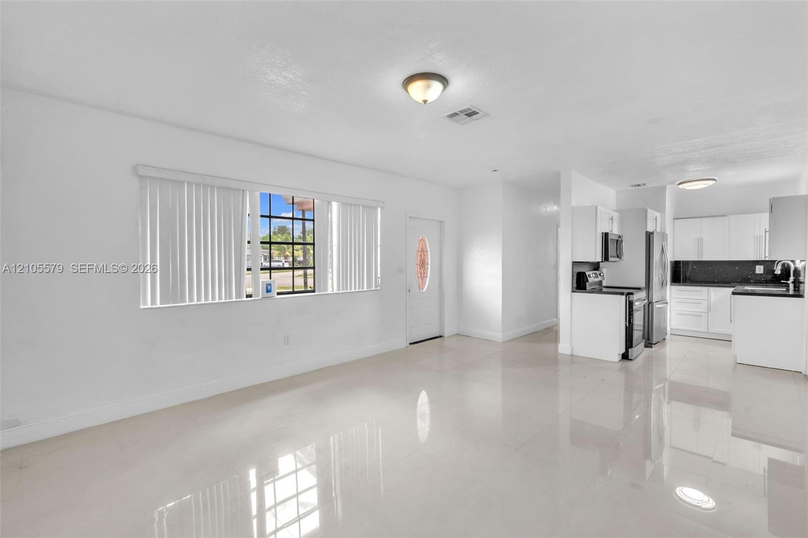 Open floor plan featuring polished tile flooring, recessed and flush-mount ceiling lighting, window with vertical blinds, and a kitchen equipped with white cabinetry and dark countertops