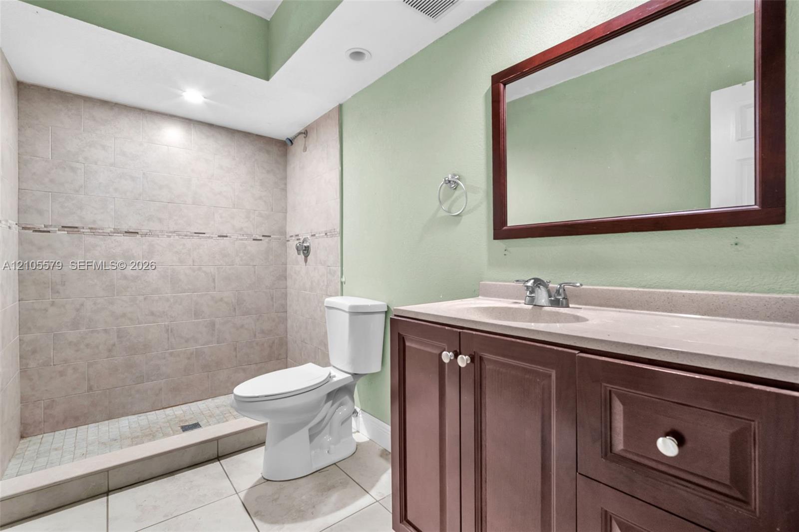 Bathroom featuring a walk-in tiled shower with an accent border, vanity with integrated stone-finish countertop, dark wood-finish cabinetry, and a framed wall-mounted mirror