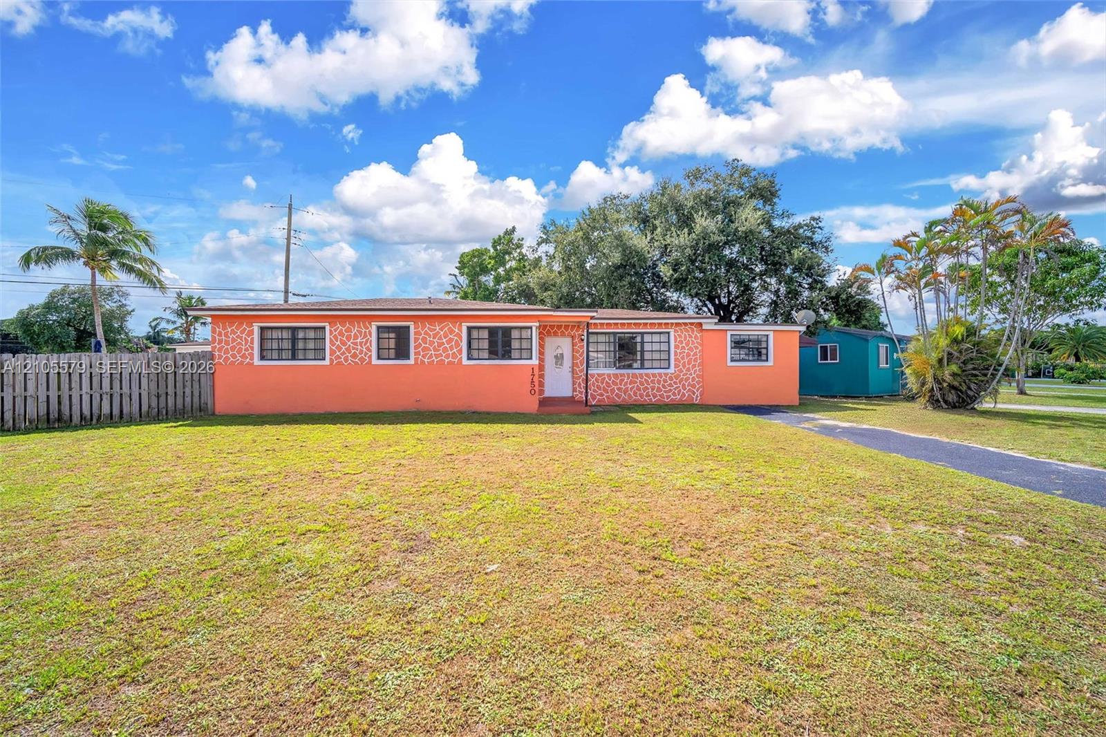 Single-story residence featuring a textured exterior facade, grid-pane windows, a paved driveway, a wood-slat perimeter fence, and mature palm trees Single-story residence featuring a textured exterior facade, grid-pane windows, a paved driveway, a wood-slat perimeter fence, and mature palm trees