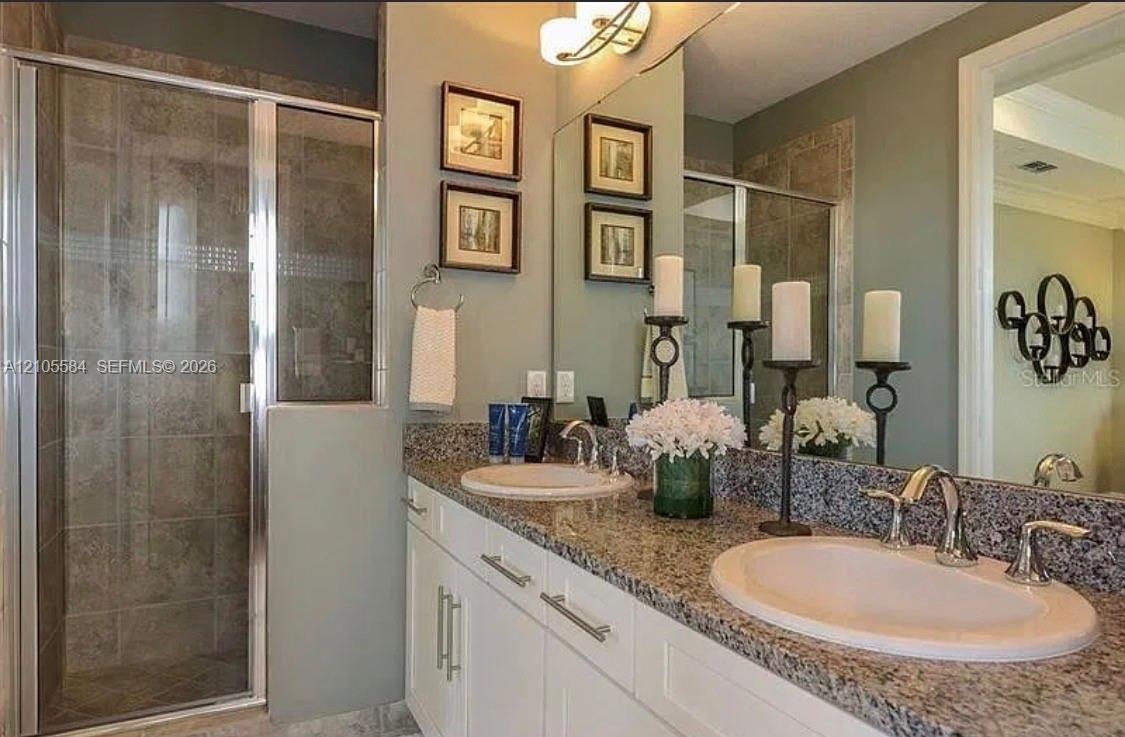 Dual-sink vanity with stone countertop, white cabinetry with brushed-metal pulls, wall-to-wall mirror, and glass-enclosed shower with tiled surround
