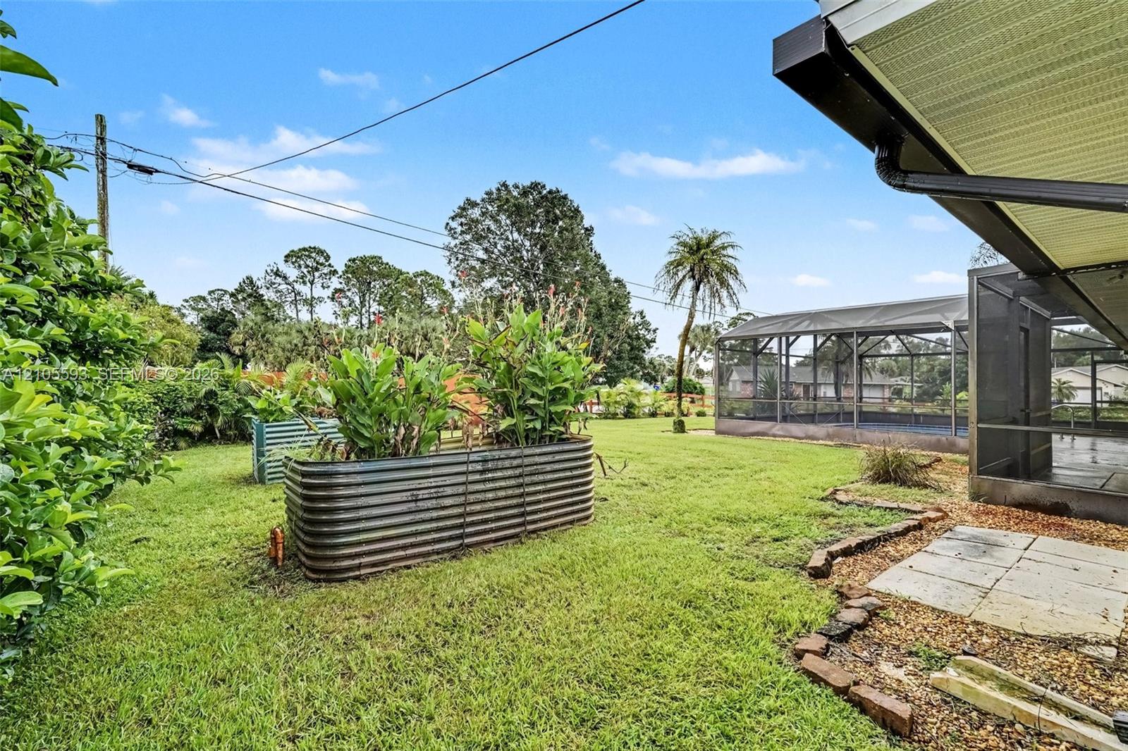 Backyard featuring a screened-in lanai, corrugated metal raised garden beds, stone paver patio area, and mature landscaping