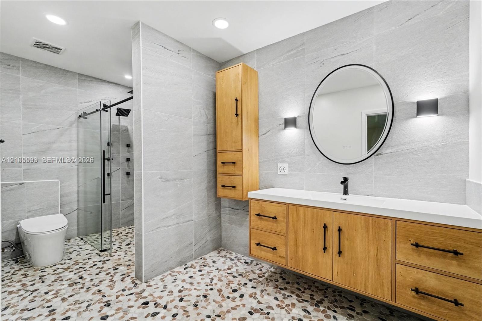 Wall-mounted wood-finish vanity with a rectangular sink, walk-in glass shower enclosure, stone-tiled walls, integrated cabinetry, and round vanity mirror