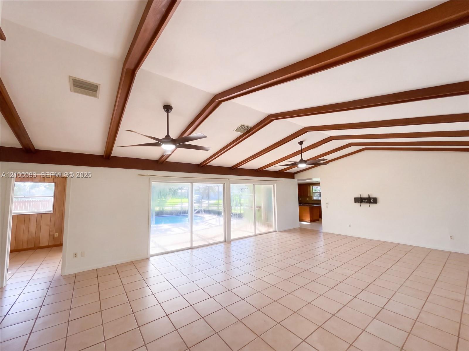 Vaulted ceiling with exposed wood beams, neutral tile flooring, dual ceiling fans with integrated lighting, and glass sliding doors
