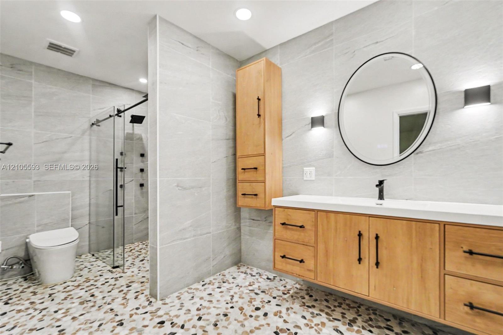 Bathroom featuring a floating vanity with wood-finish cabinetry, a white countertop, a walk-in shower with a glass enclosure, wall-to-wall stone-tiled surfaces, and river-stone flooring