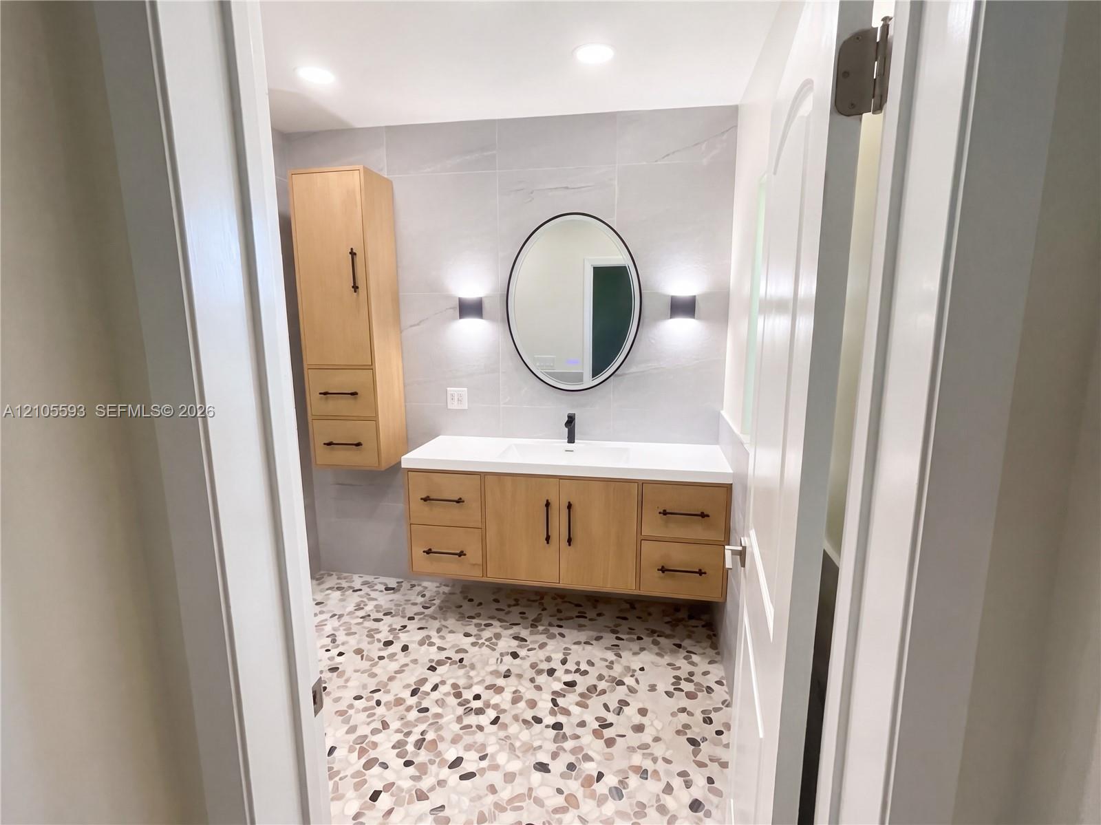 Floating wood-finish vanity with matte black hardware, integrated rectangular sink, wall-mounted oval mirror, light-toned stone-patterned wall tile, and pebble-textured floor tile