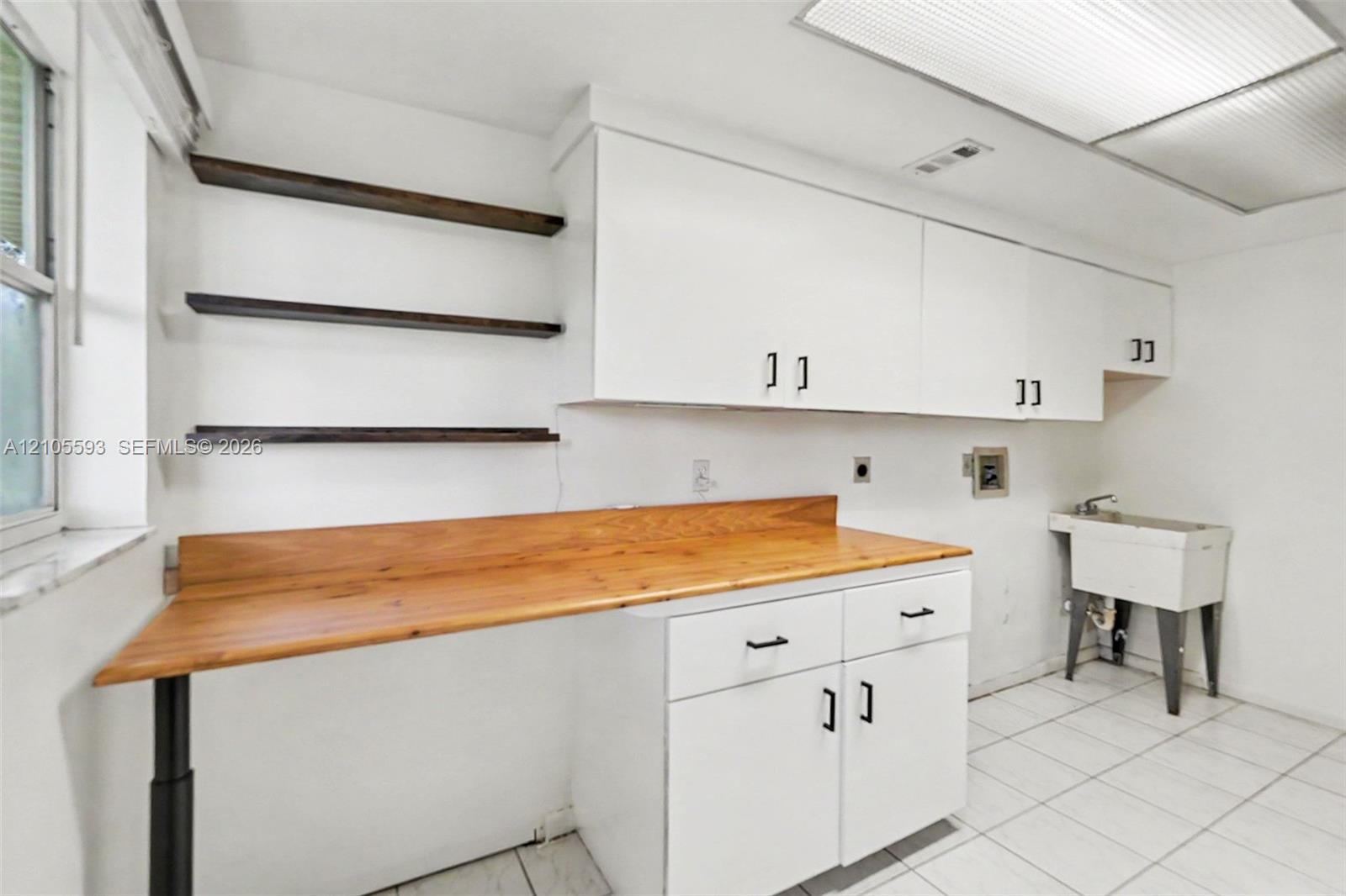 Laundry area featuring white cabinetry, a wood-finish countertop workspace, open shelving, a utility sink, and tile flooring
