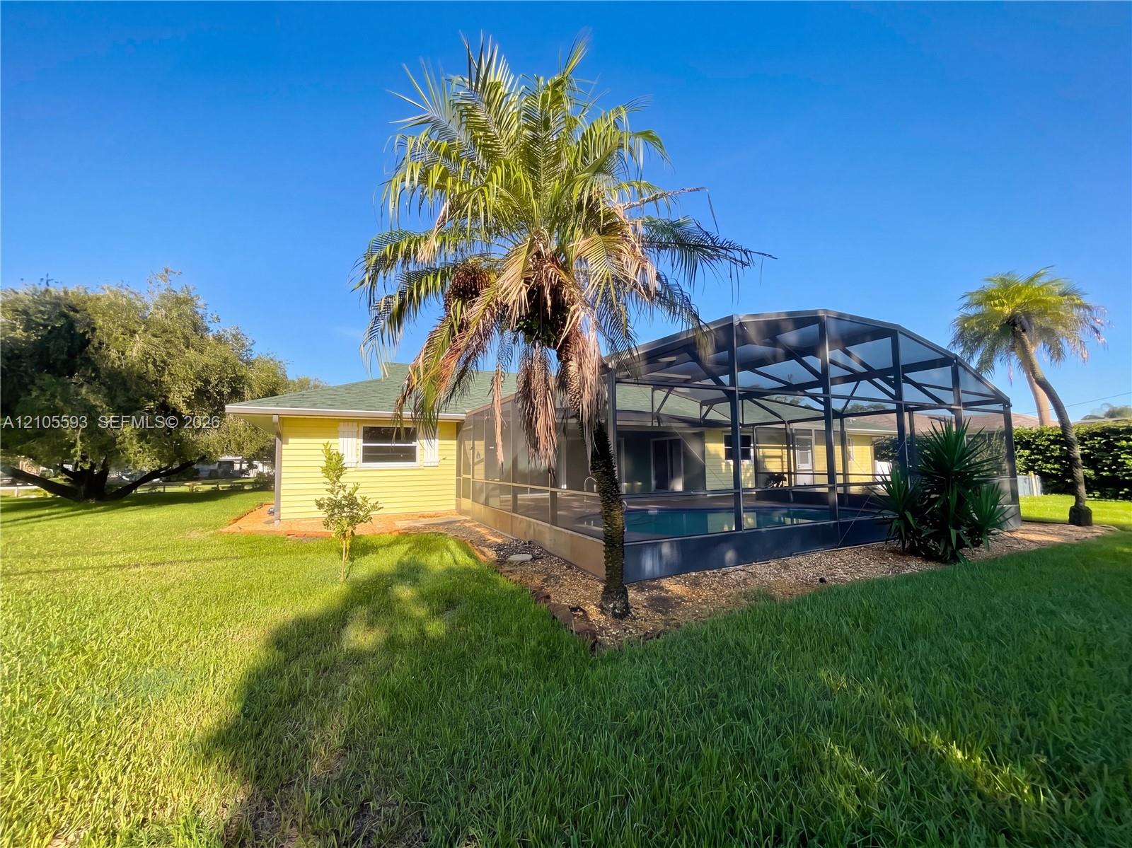 Exterior of the residence featuring light yellow lap siding, an enclosed screened-in pool, mature palm trees, and an expansive lawn